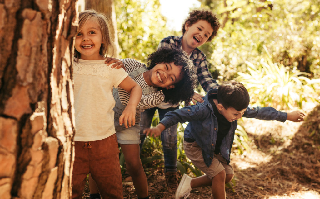 Four children peek out from behind a tree in a forest, laughing and playing