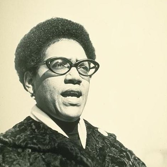 A photo of poet and activist Audre Lorde, a Black woman with short hair wearing glasses and a dark-colored coat