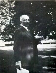 A black and white photo of a Black man in a dark -colored suit standing in front of a grassy field and a tree