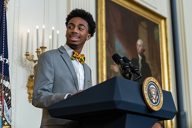 A photograph of a black man in a gray suit with a colorful bowtie standing in the White House in front of a presidential podium